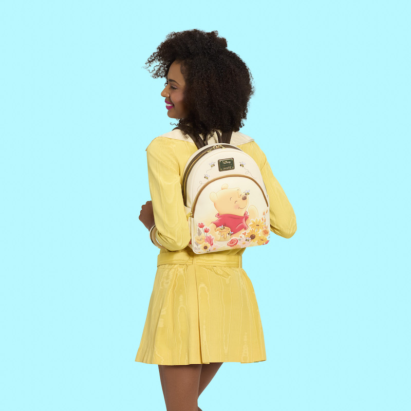 Woman wearing a yellow dress, facing away from camera. She wears the Loungefly Winnie the Pooh mini backpack on her back. She stands against a blue background. 
